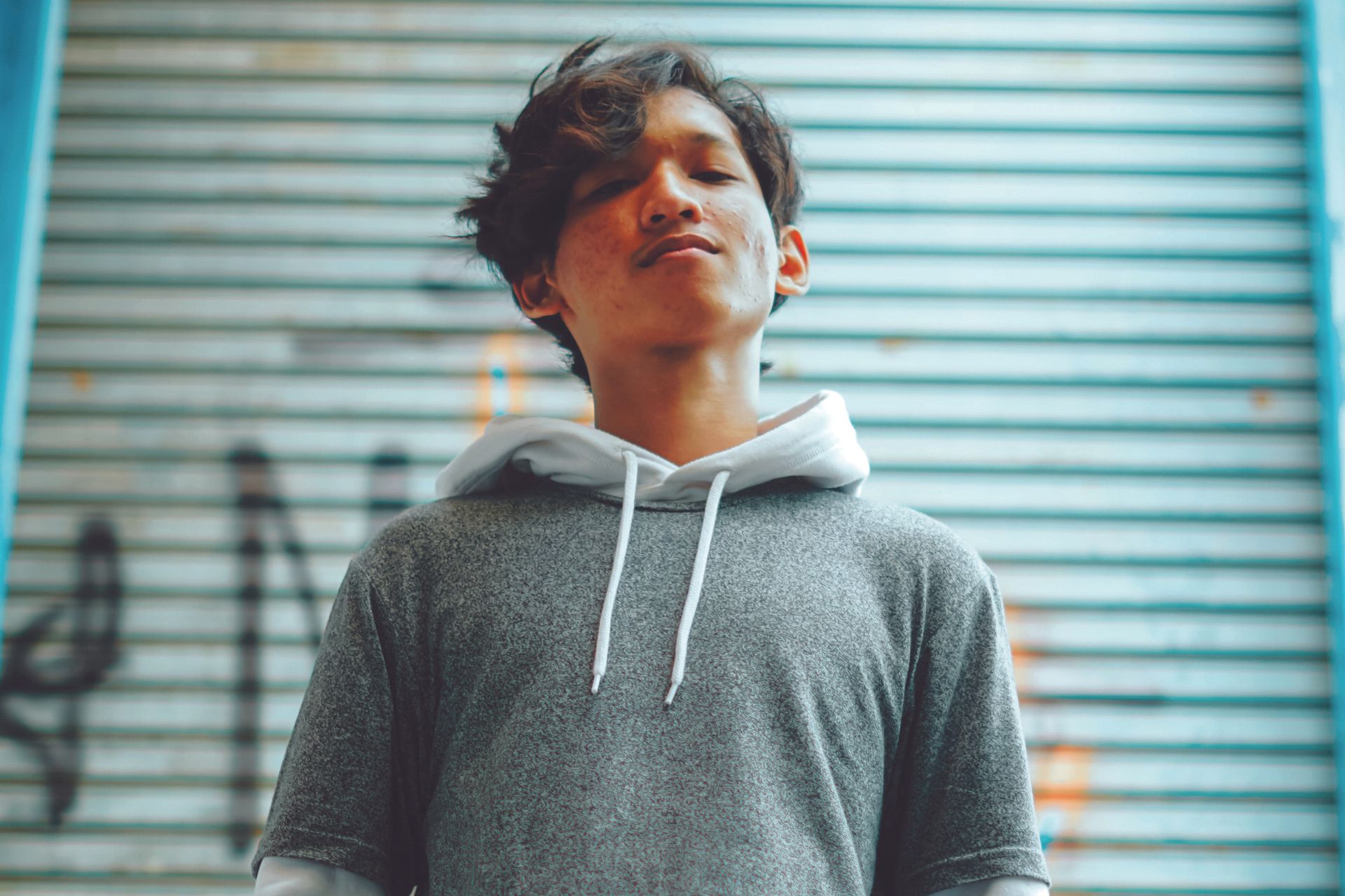 A young person outside facing directly at the camera, with a graffiti-covered metal wall in the background.