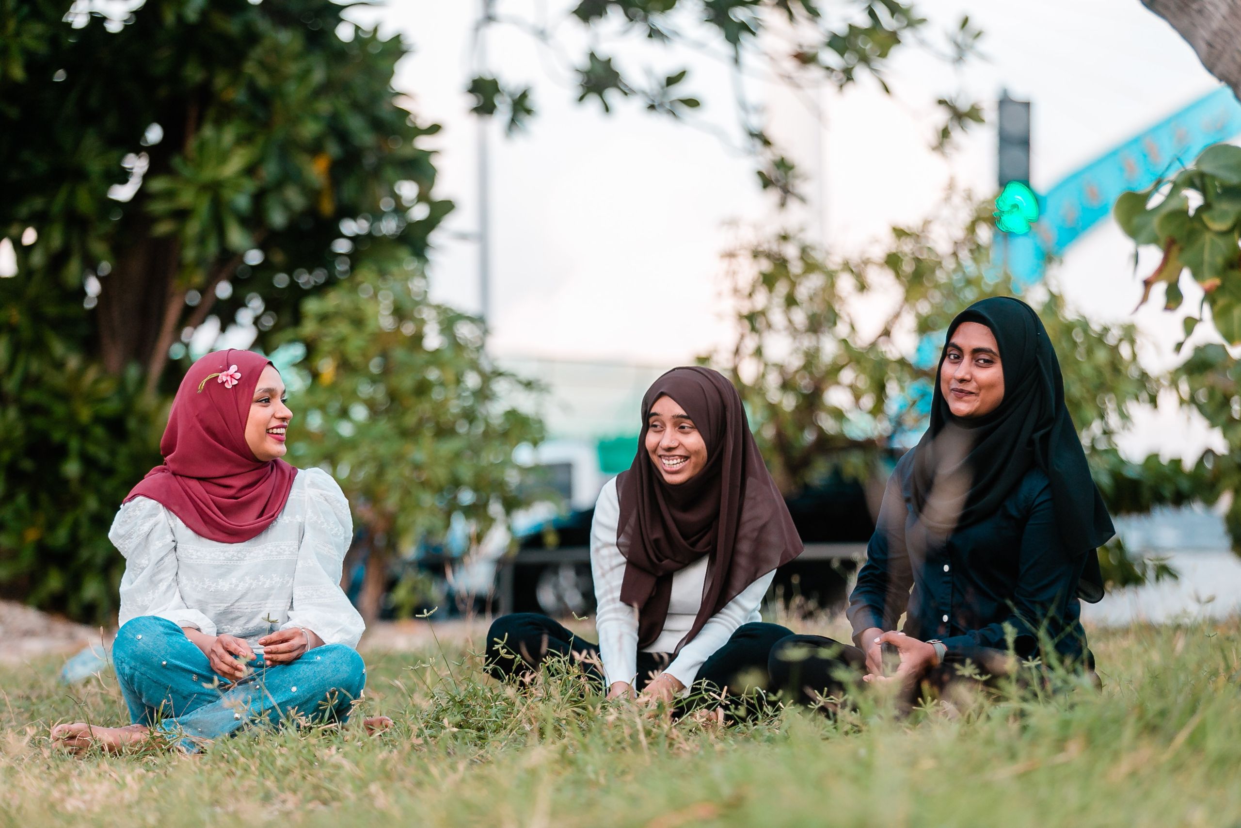 Three newcomer youth sitting on the grass in a park, wearing hijabs